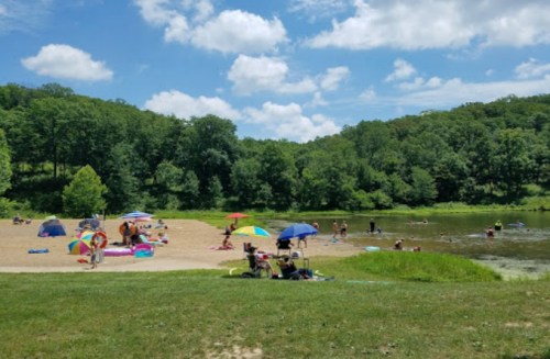 A sunny beach scene with people relaxing on the sand, swimming in the water, and enjoying colorful umbrellas and floats.