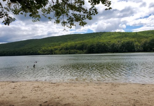 A serene lake surrounded by green hills under a cloudy sky, with a sandy beach in the foreground.