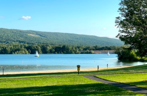 A serene lake scene with sailboats, green grass, and mountains in the background under a clear blue sky.