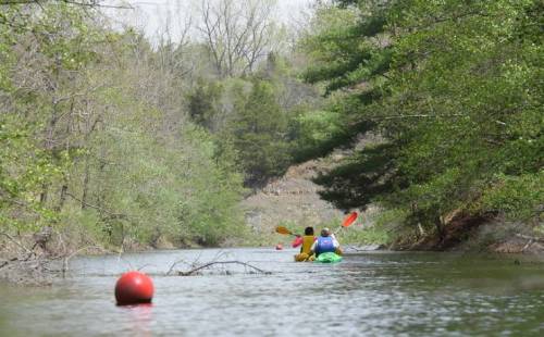 Two kayakers paddle down a serene, tree-lined river with a red buoy in the foreground.