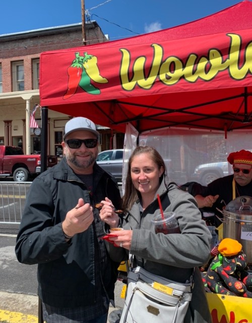 A man and woman smile while holding cups of food at a colorful festival booth with a red canopy.