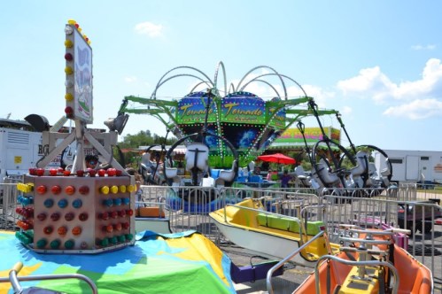 Colorful carnival rides and games under a clear blue sky, with vibrant decorations and seating areas in the background.