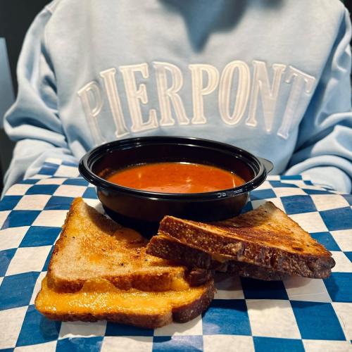 A bowl of tomato soup with grilled cheese sandwiches on a checkered paper, worn by a person in a "PIERPONT" sweatshirt.