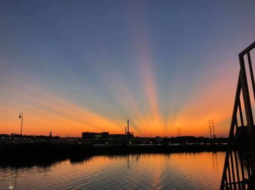 A vibrant sunset with rays of light spreading across the sky, reflecting on calm water. Silhouettes of buildings in the background.