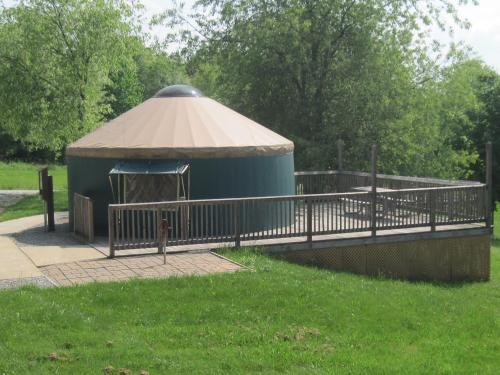 A round, green yurt with a tan roof, surrounded by a wooden deck and grassy area, under a clear blue sky.