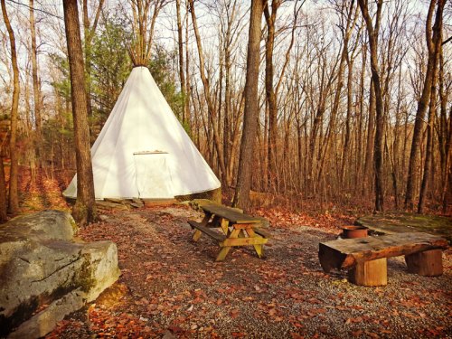 A white teepee surrounded by trees, with picnic tables and a stone fire pit on a leaf-covered ground.