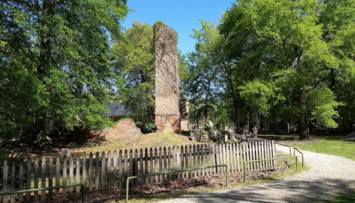 A brick chimney stands among trees, with a gravel path and a wooden fence in the foreground.