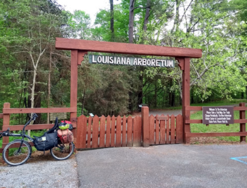 A wooden entrance sign for the Louisiana Arboretum, with a bicycle parked nearby amidst lush greenery.