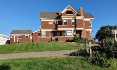 A large, two-story red brick house with a sloped roof, surrounded by green grass and blue sky.