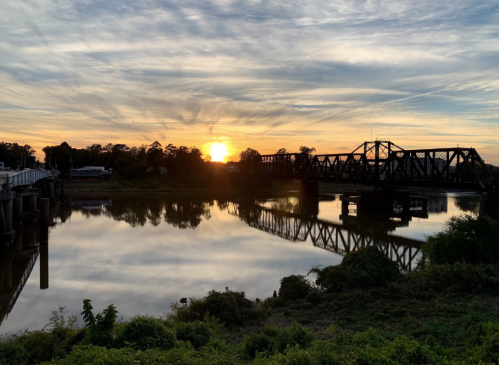 Sunset over a river with a bridge reflected in the water, surrounded by trees and a cloudy sky.