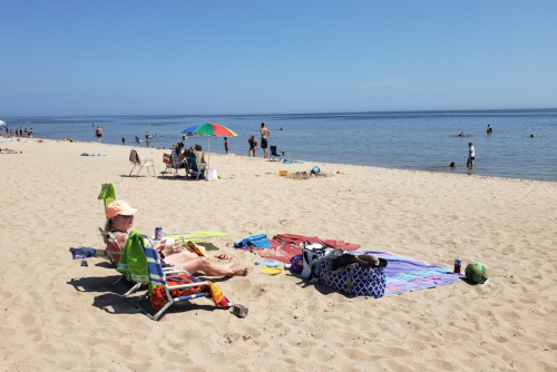 A sunny beach scene with people relaxing, swimming, and enjoying the water, along with colorful umbrellas and towels.