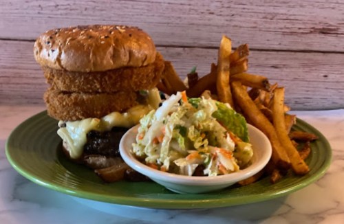 A towering burger with cheese and onion rings, served with fries and a side of coleslaw on a green plate.
