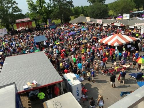 A large crowd gathers at an outdoor festival with colorful tents and a stage in the background.
