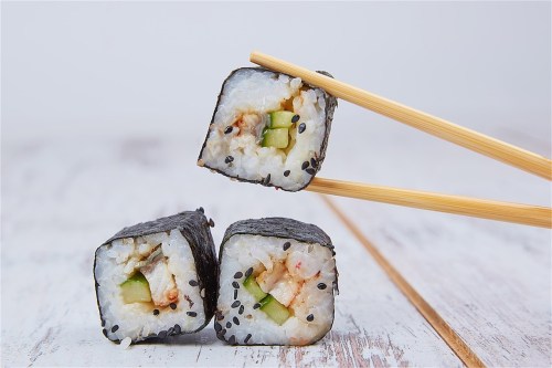 Three pieces of sushi with shrimp and cucumber, held by chopsticks, on a wooden surface.
