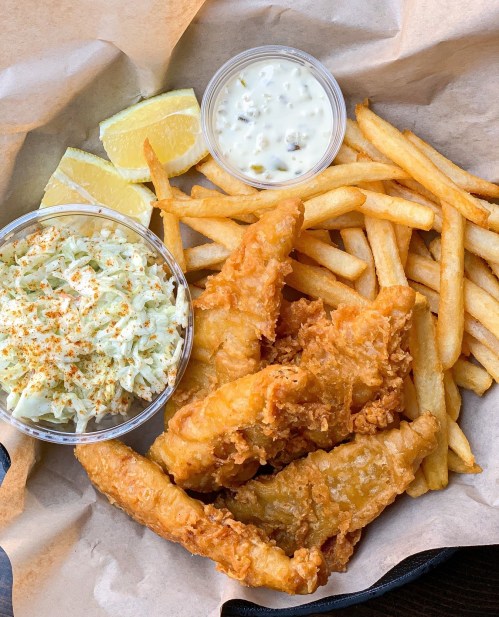 A serving of fried fish fillets with fries, coleslaw, lemon wedges, and a side of tartar sauce on parchment paper.