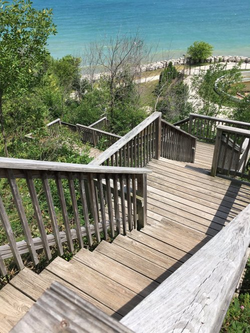 Wooden stairs lead down to a beach with clear blue water, surrounded by greenery and trees.