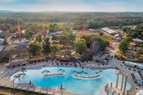 Aerial view of a water park with a pool, palm trees, and a roller coaster in a lush green landscape.
