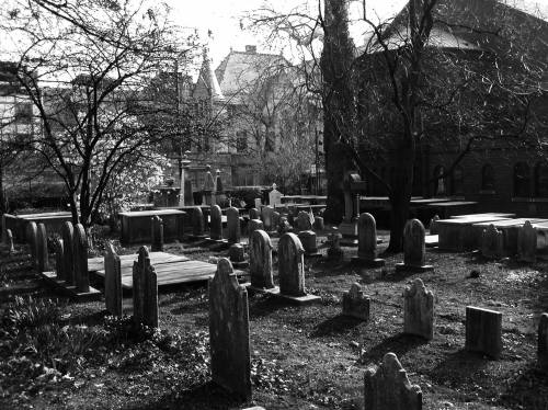A black and white image of a cemetery with various gravestones, surrounded by trees and historic buildings.