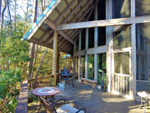 A wooden deck with a table and chairs, surrounded by trees and featuring large windows in a rustic cabin.