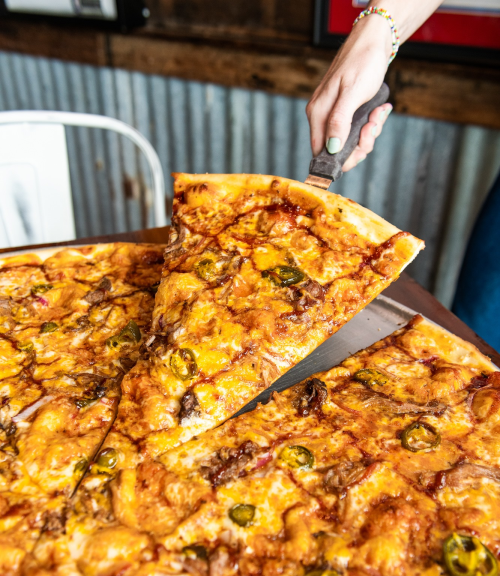 A hand lifts a slice of pizza topped with cheese, jalapeños, and other toppings from a large pizza on a wooden table.