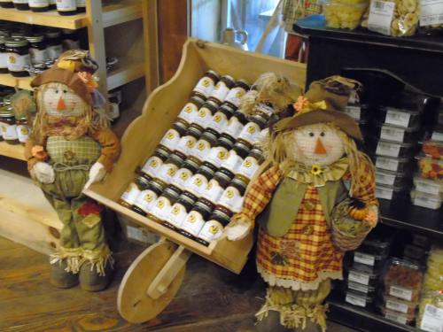 Two scarecrow decorations stand beside a wooden wheelbarrow filled with jars, set in a cozy shop interior.