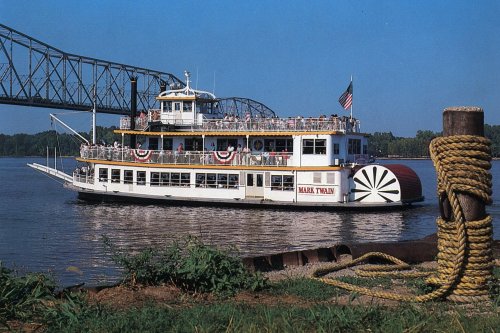 A riverboat named "Mark Twain" sails on a calm river, with a bridge in the background and a rope tied to a post nearby.