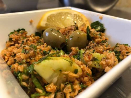 A close-up of a bowl of bulgur salad with herbs, olives, and lemon slices, garnished with fresh greens.