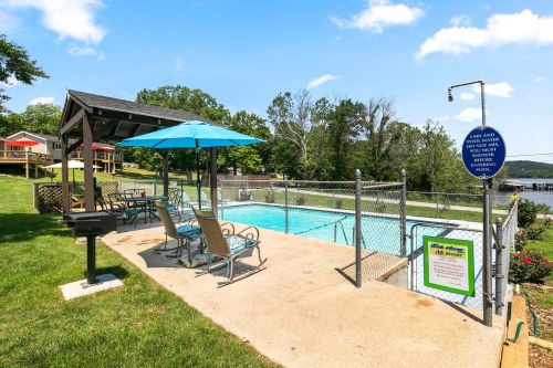 A fenced pool area with lounge chairs, an umbrella, and a grill, surrounded by greenery and a view of nearby houses.