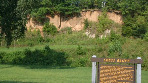 A grassy area with a sign reading "Loess Bluff" in the foreground, and a steep, sandy bluff in the background.