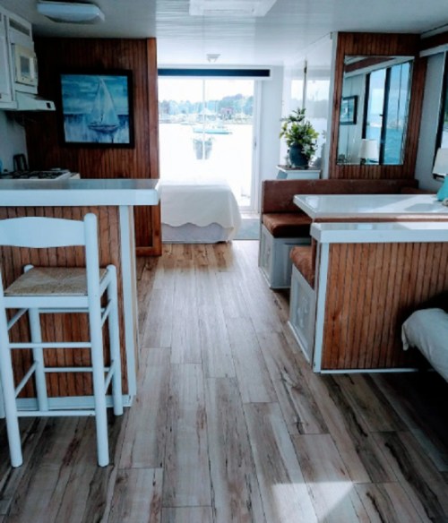 Interior of a cozy boat cabin featuring a kitchenette, dining area, and a bed with large windows showing a view outside.