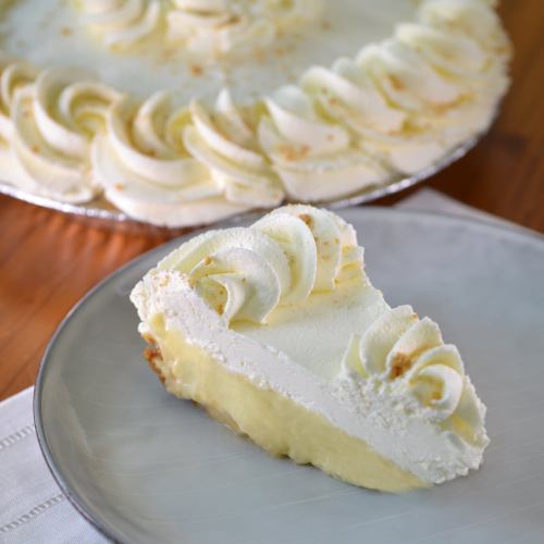 A slice of creamy pie topped with whipped cream, served on a plate, with the whole pie in the background.