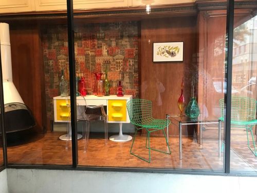 A modern interior display featuring colorful glass vases, a white table, and green wire chairs behind a glass window.