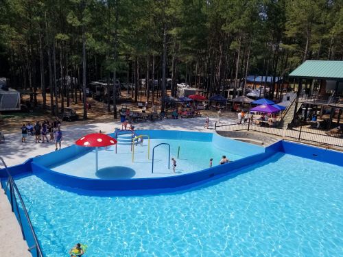 A bright blue pool with water features, surrounded by trees and people enjoying a sunny day at a recreational area.
