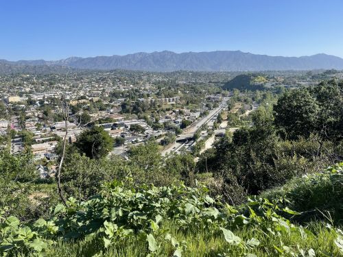 A panoramic view of a city with mountains in the background, greenery in the foreground, and a clear blue sky.