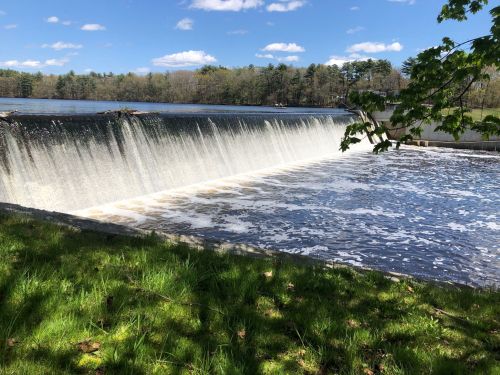 A serene view of a waterfall cascading over a dam into a calm lake, surrounded by greenery and blue skies.