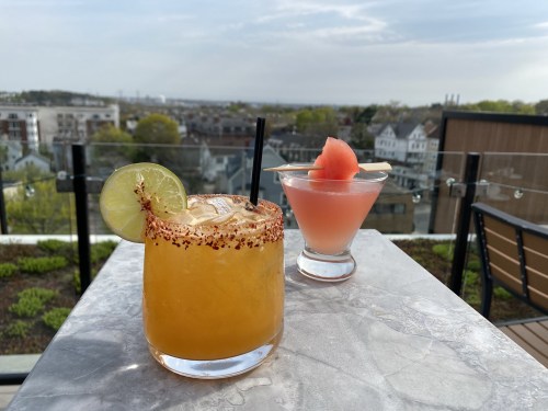 Two cocktails on a marble table with a cityscape in the background; one is garnished with a lime and chili rim.
