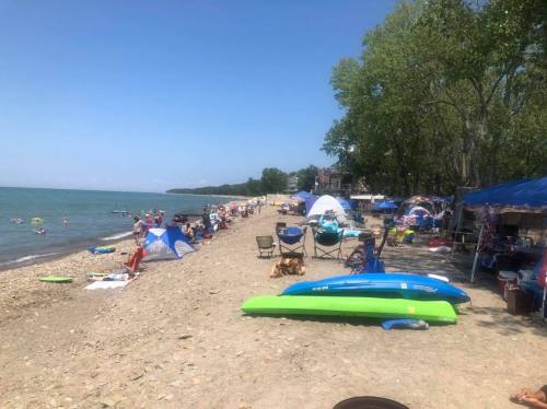 A sunny beach scene with people relaxing, tents set up, and kayaks along the shore by a calm lake.