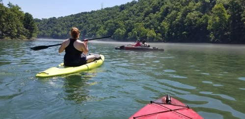 A person paddles a yellow kayak on a calm lake, with another kayak in the distance surrounded by lush greenery.