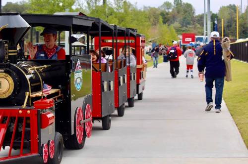 A small train with passengers rides along a path, while people walk nearby in a park setting.