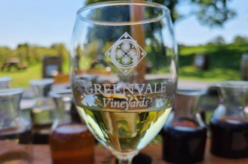 A glass of white wine with the Greenvale Vineyards logo, surrounded by small carafes of various wines outdoors.