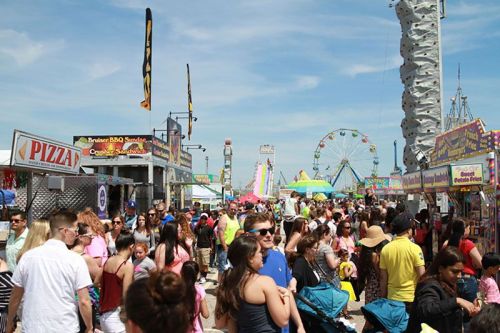 A bustling fairground scene with crowds, food stalls, and amusement rides under a clear blue sky.