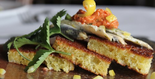 A plated dish featuring grilled fish on cornbread, topped with sauce and garnished with herbs and diced vegetables.