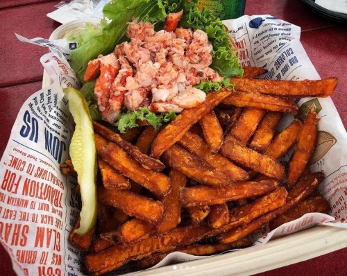 A basket of sweet potato fries with a side of lobster salad on lettuce and a pickle.
