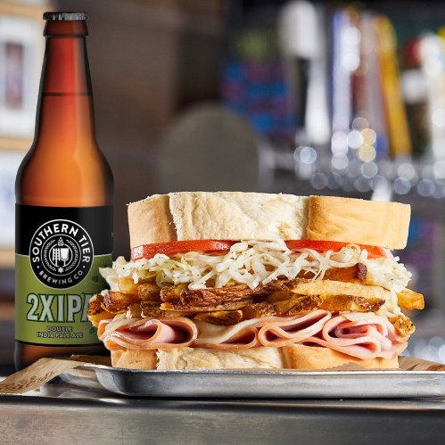 A close-up of a hearty sandwich with ham, lettuce, and fries, next to a bottle of Southern Tier 2X IPA beer.
