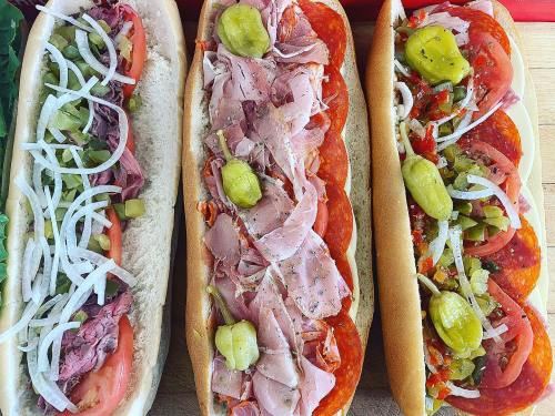Three sub sandwiches with various meats, toppings, and peppers on hoagie rolls, arranged on a wooden surface.