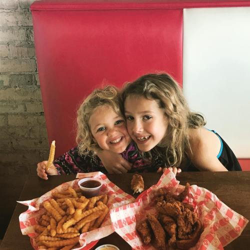 Two smiling girls sit at a table with plates of fried chicken and crinkle-cut fries, enjoying their meal together.