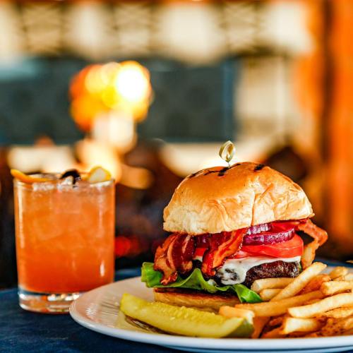 A juicy burger with bacon, lettuce, and tomato, served with fries and a cocktail on a table.