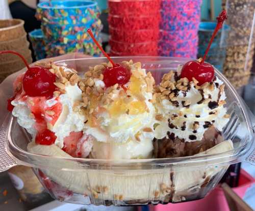 A colorful ice cream sundae with three scoops, topped with whipped cream, nuts, and cherries, served in a clear container.