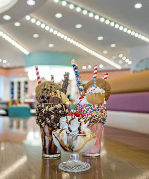 Three colorful ice cream sundaes with toppings, including sprinkles, cookies, and cherries, set in a bright café.