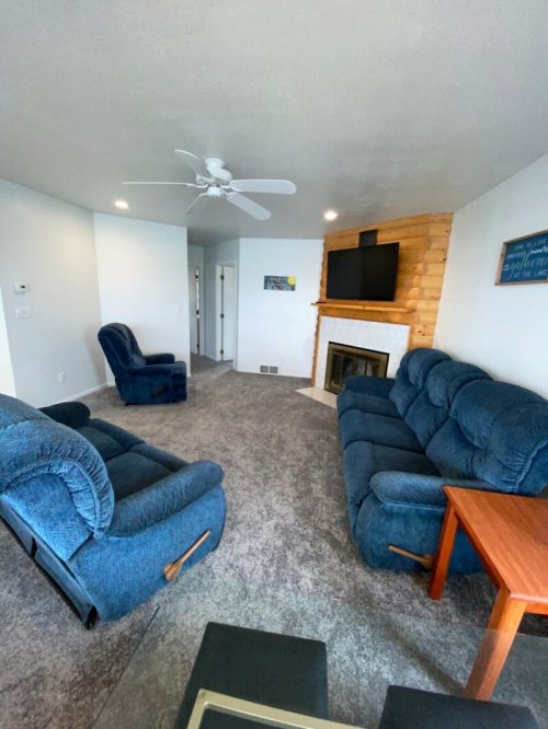 Cozy living room with blue sofas, a recliner, a fireplace, and a TV on a wooden wall. Light walls and carpeted floor.